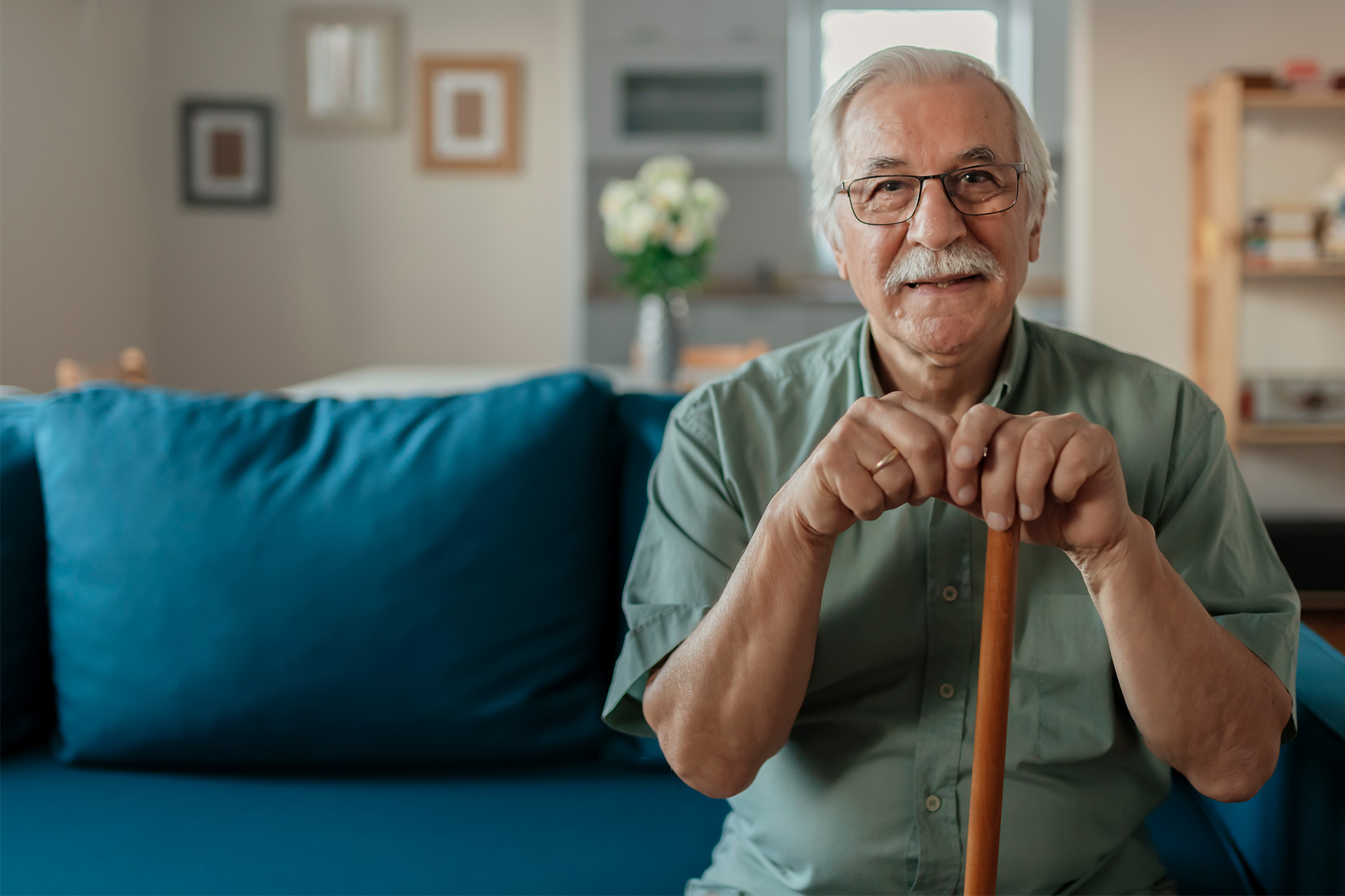 Lächelnder älterer Mann mit Brille und Krückstock sitzt auf einem blauen Sofa in einem gemütlichen Wohnzimmer.