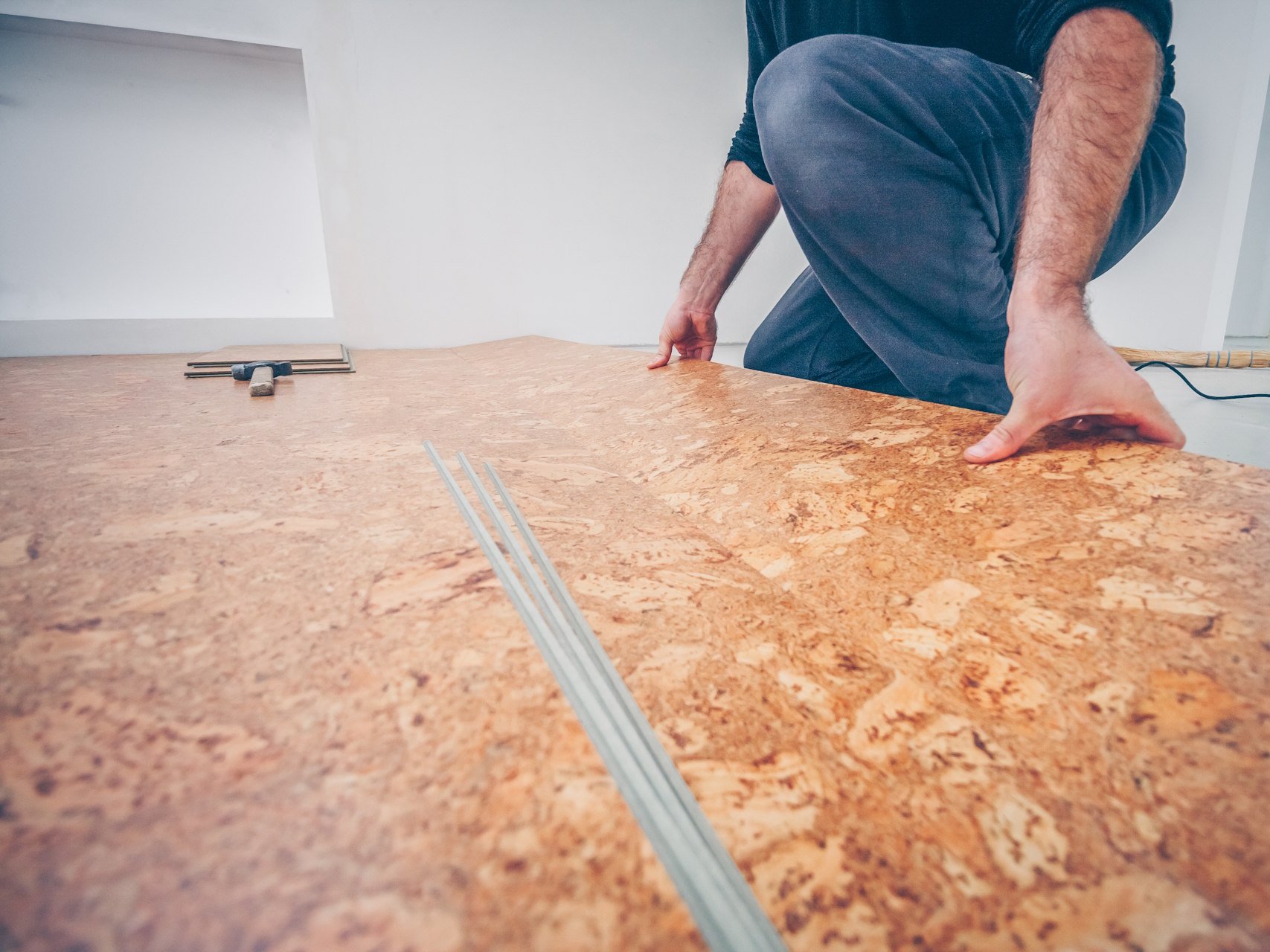 Lassen Sie Ihren Korkboden richtig verlegen von unserem Fachbetrieb in Gronau, um die optimale Elastizität und Langlebigkeit dieses Naturmaterials voll auszuschöpfen. Wir garantieren Ihnen eine präzise Ausführung und die fachgerechte Vorbereitung des Untergrunds für ein perfektes, wohngesundes Ergebnis in jedem Raum.  Bildbeschreibung Eine bodennahe Aufnahme zeigt eine Person in kniender Haltung, die eine großformatige Korkplatte verlegt. Die Hände der Person drücken die Platte flach auf den Boden, um sie bündig an die bereits liegende Fläche anzuschließen. Der Korkboden weist eine markante, lebendige Struktur in verschiedenen Brauntönen auf; im unscharfen Hintergrund sind Werkzeuge wie ein Hammer erkennbar, die auf eine professionelle Montage hindeuten.