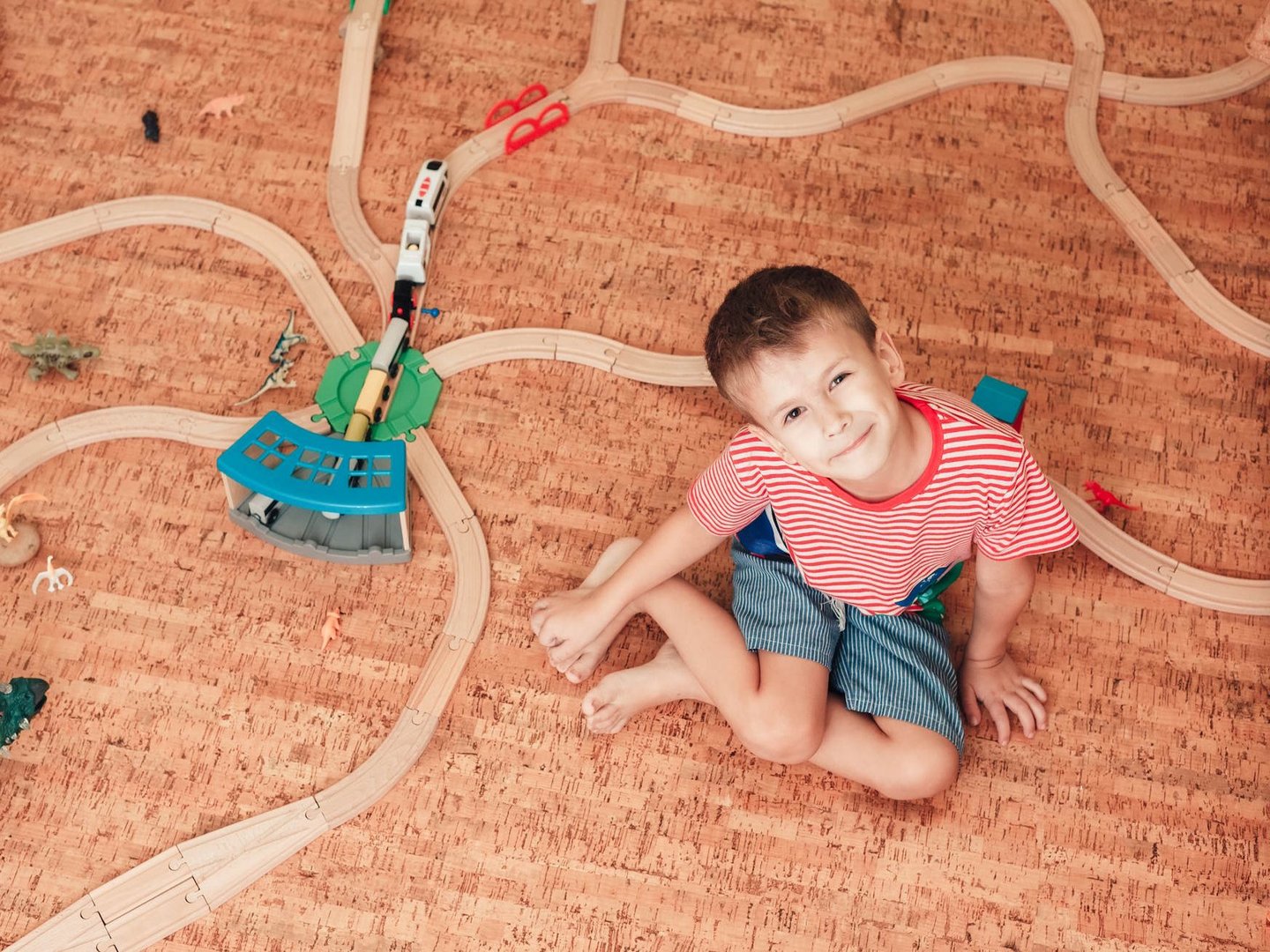 Nutzen Sie die natürlichen Vorteile von Korkboden bei Ihrem Fachhändler in Gronau und schaffen Sie eine wohngesunde, fußwarme Umgebung für Ihre gesamte Familie. Dank seiner elastischen und schalldämmenden Eigenschaften ist Kork der ideale Bodenbelag für Kinderzimmer, der höchsten Komfort mit nachhaltigem Umweltschutz verbindet.  Bildbeschreibung Die Draufsicht zeigt ein Kind, das lächelnd auf einem hellbraunen Korkboden mit charakteristischer, feinkörniger Naturstruktur sitzt. Um das Kind herum ist eine hölzerne Spielzeugeisenbahn auf den Korkplatten aufgebaut, ergänzt durch kleine Spielfiguren. Das warme Erscheinungsbild des Bodens unterstreicht die gemütliche und sichere Atmosphäre einer Spielumgebung im Kinderzimmer.