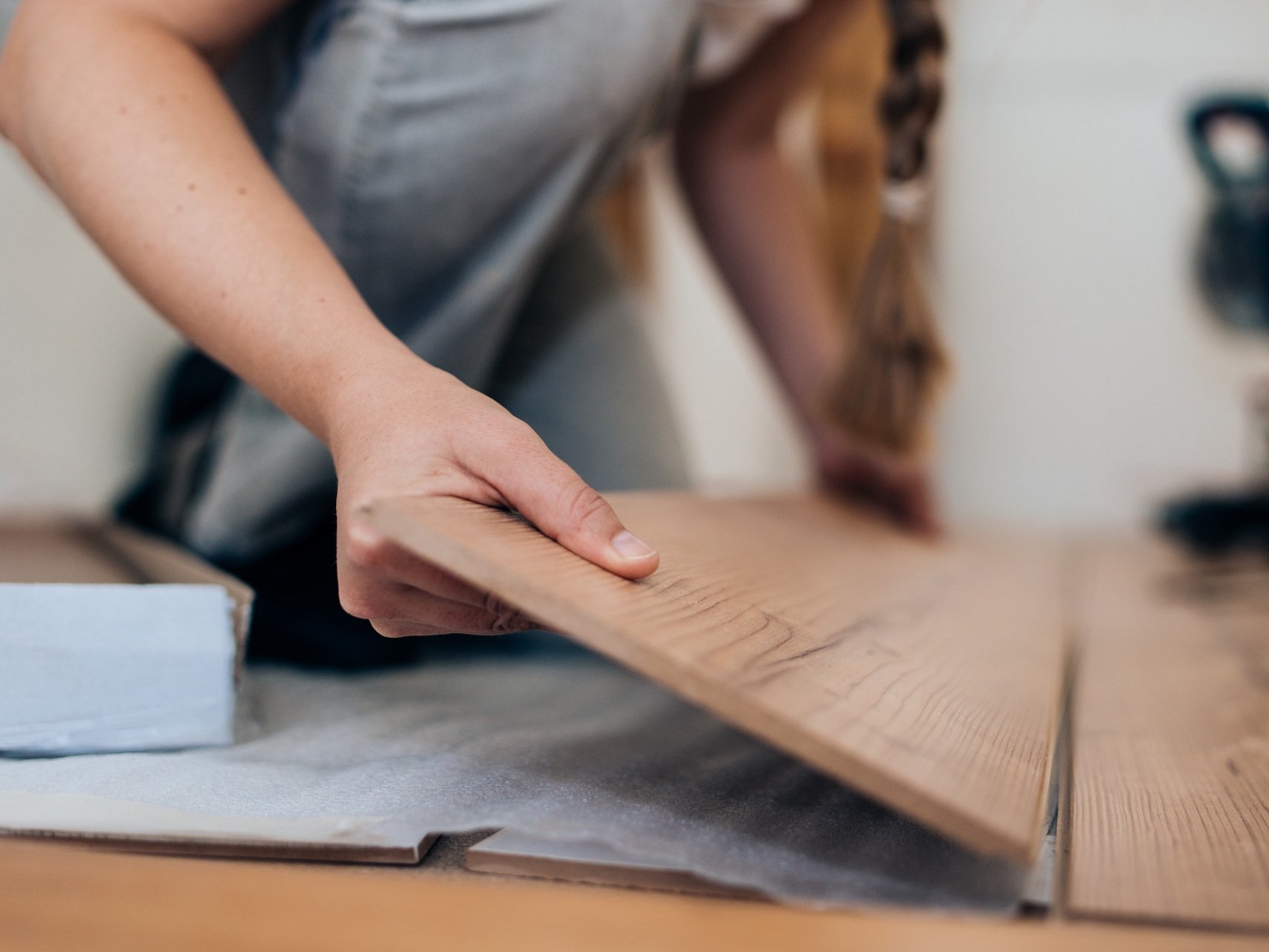 Überlassen Sie das Laminatboden-Verlegen unserem erfahrenen Fachbetrieb in Gronau, um ein präzises und langlebiges Ergebnis in Ihren Wohnräumen zu garantieren. Wir sorgen für eine fachgerechte Verlegung inklusive Trittschalldämmung, damit Sie lange Freude an Ihrem neuen, makellosen Bodenbelag haben.  Bildbeschreibung Eine Nahaufnahme zeigt eine Person in Arbeitskleidung, die kniend eine Paneele Laminatboden mit natürlicher Holzmaserung verlegt. Die Hände halten das Brett in einem schrägen Winkel an die bereits verlegte Fläche an, um es in das Klick-System einzurasten. Unter dem Bodenbelag ist eine helle Trittschalldämmung sichtbar; der Fokus liegt scharf auf der Hand und der Kante des Laminatbretts, während der Hintergrund unscharf gehalten ist.