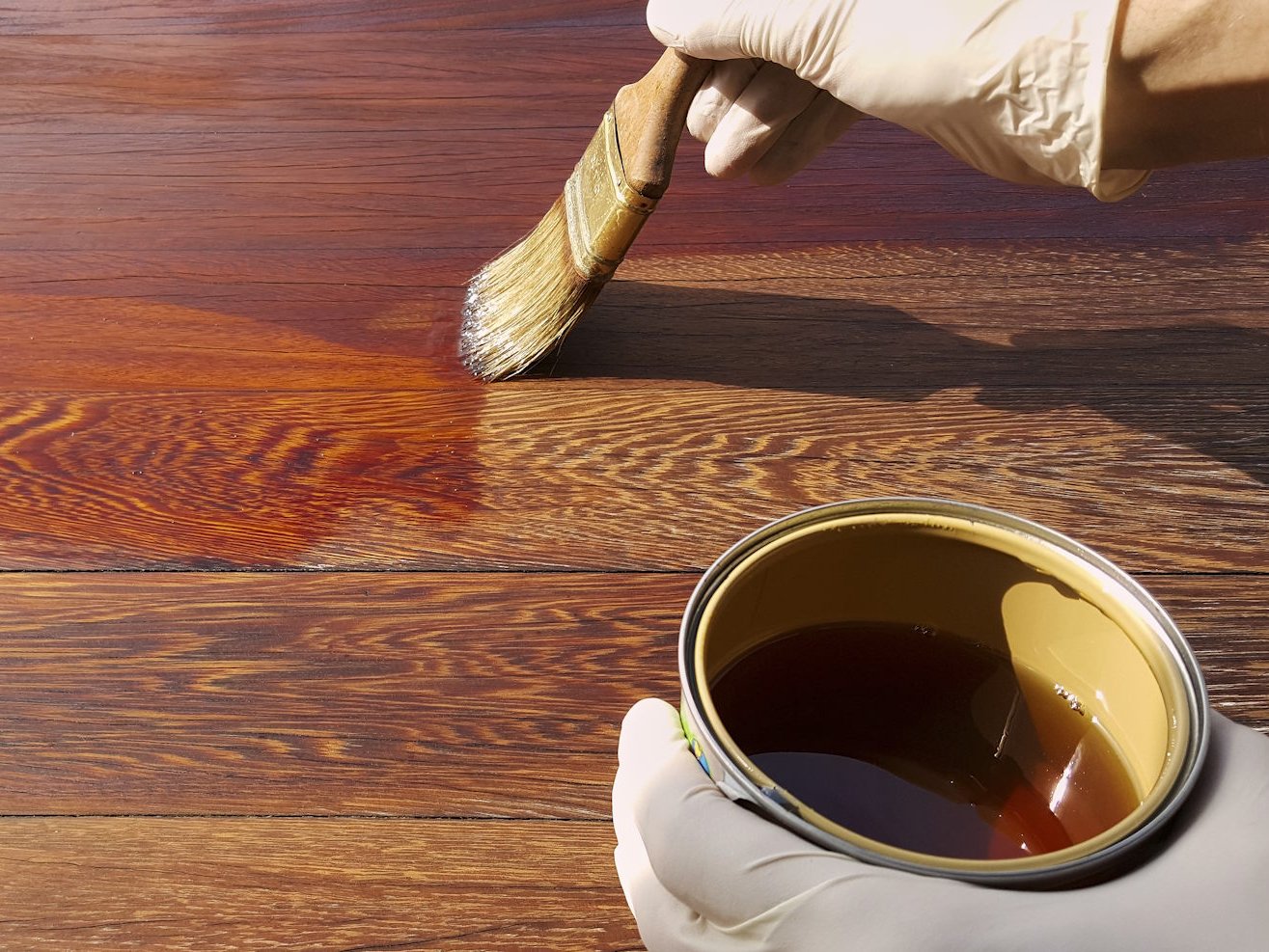 Vertrauen Sie auf professionelle Pflegeprodukte und Expertenrat von Ihrem Fachhandel in Gronau, um die natürliche Schönheit Ihrer Massivholzdielen über Jahrzehnte zu erhalten. Durch regelmäßiges Ölen und die richtige Oberflächenbehandlung schützen Sie Ihren wertvollen Echtholzboden nachhaltig vor äußeren Einflüssen und bewahren seine einzigartige Ausstrahlung.  Bildbeschreibung Eine Detailaufnahme zeigt eine Person mit weißen Schutzhandschuhen, die mit einem breiten Lasurpinsel eine dunkle, bernsteinfarbene Pflegeflüssigkeit oder Öl auf eine hölzerne Dielenoberfläche aufträgt. In der unteren Bildhälfte ist eine geöffnete Metalldose mit dem flüssigen Pflegemittel zu sehen. Der Kontrast zwischen der behandelten, glänzenden Holzfläche und dem noch unbehandelten, matten Bereich betont die intensive Maserung und die warme Färbung des Holzes.