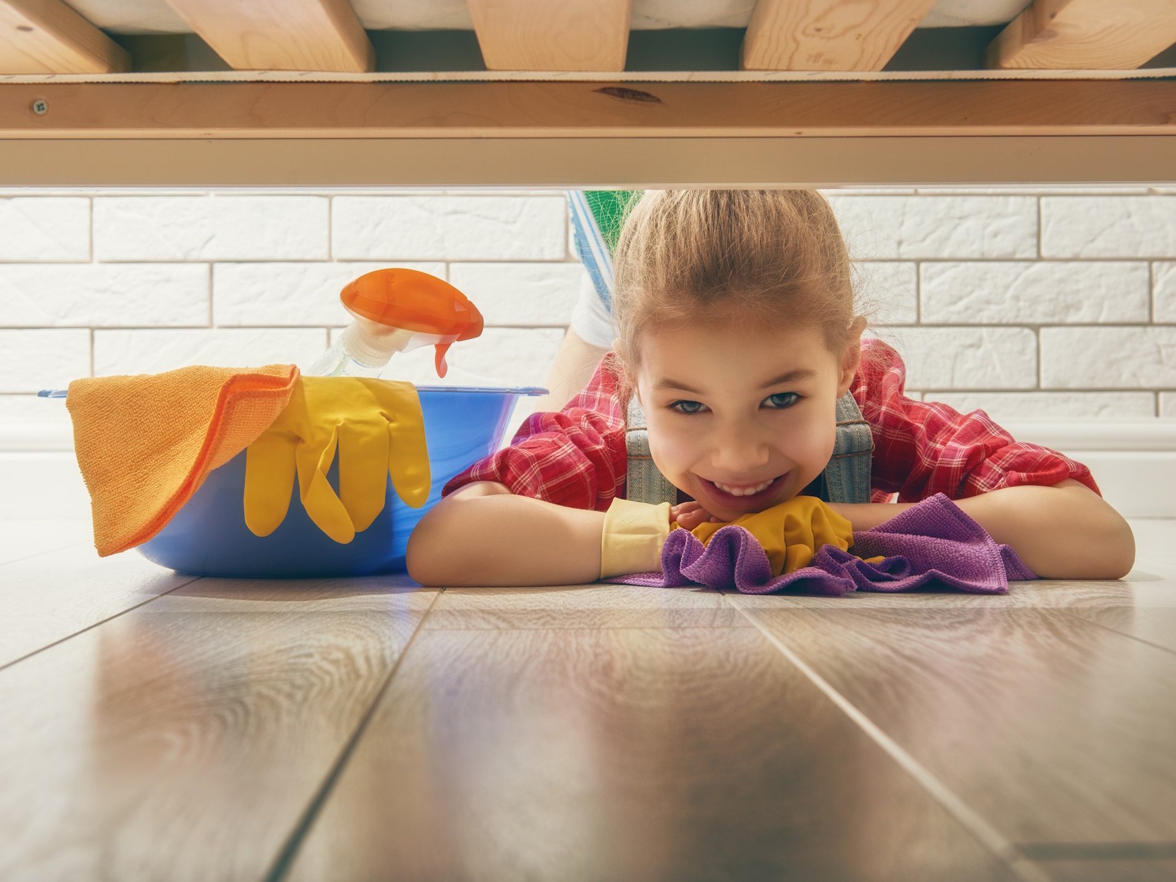 Sorgen Sie für langanhaltenden Glanz mit spezialisierten Pflegemitteln für Parkett, Laminat & Co, die Sie direkt in Gronau beziehen können. Die regelmäßige Reinigung mit abgestimmten Produkten schützt die Oberfläche Ihres Holzfussbodens effektiv vor Abnutzung und erhält die natürliche Schönheit über viele Jahre.  Beschreibung: Ein lächelndes Mädchen liegt bäuchlings auf einem hellen Holzfußboden und schaut unter einem Möbelstück hervor. Neben ihr befinden sich Reinigungsutensilien wie ein blauer Eimer, gelbe Gummihandschuhe, ein violettes Mikrofasertuch und eine Sprühflasche. Das Bild vermittelt eine positive Atmosphäre bei der Bodenpflege und Reinigung im häuslichen Umfeld.