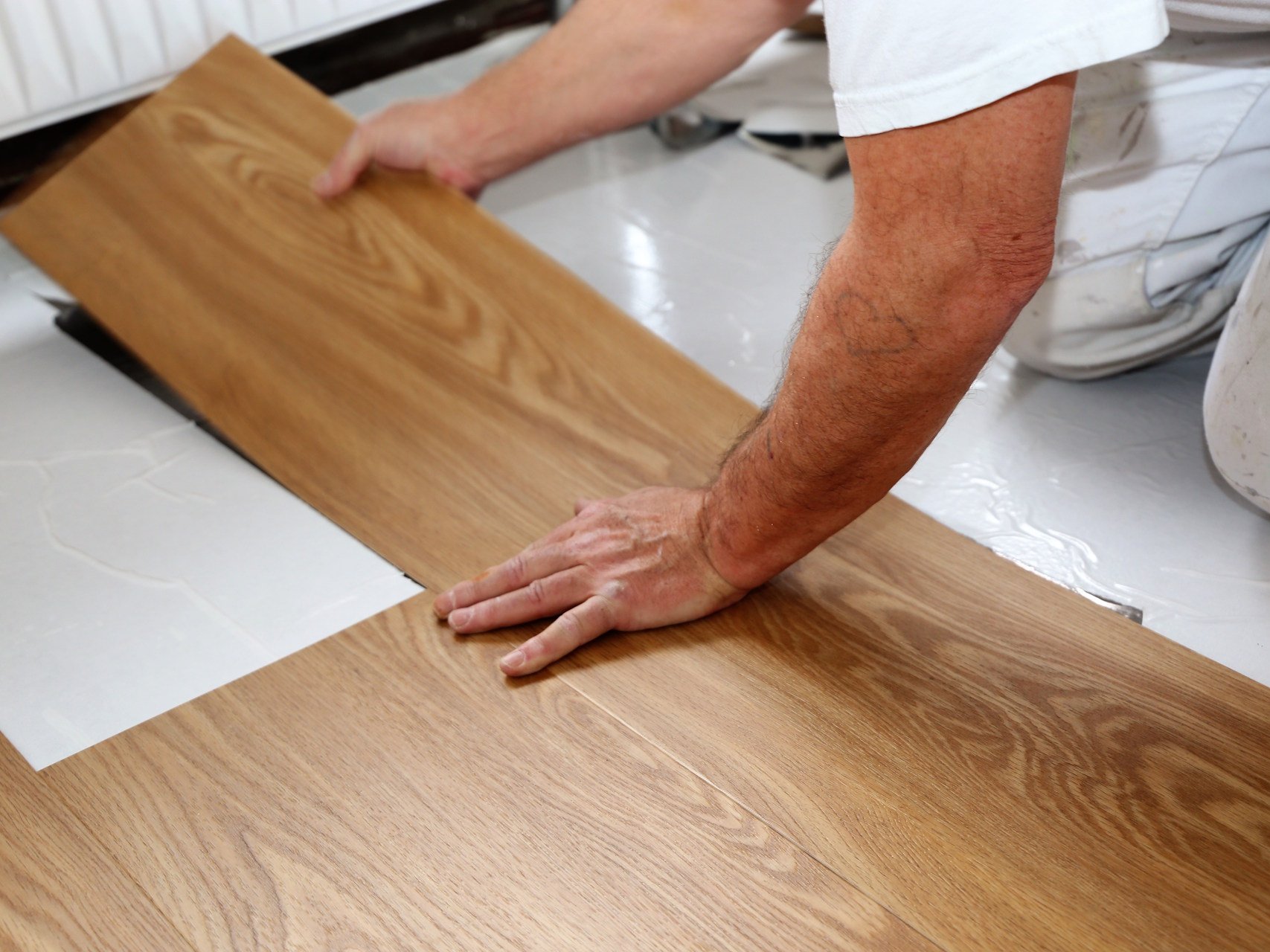 Lassen Sie Ihr Designvinyl professionell verlegen von unserem erfahrenen Fachbetrieb in Gronau, um ein perfektes und langlebiges Bodenbild zu garantieren. Wir sorgen für eine fachgerechte Untergrundvorbereitung und präzise Klick-Verlegung, damit Ihr neuer Vinylboden auch bei hoher Beanspruchung seine erstklassige Optik behält.  Bildbeschreibung Die Nahaufnahme zeigt eine Person in weißer Arbeitskleidung, die eine Vinyl-Diele in warmer Eichenoptik auf einem weißen Untergrund verlegt. Die Hände führen das Paneel präzise in das Klick-System der bereits liegenden Bodenfläche ein. Der Fokus liegt scharf auf der Holzstruktur des Bodenbelags und der ansetzenden Hand, während der Hintergrund mit einem Heizkörper leicht in Unschärfe verschwimmt.